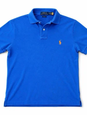 Blå pikétröja från Polo Ralph Lauren - Klassisk blå pikétröja från Polo Ralph Lauren med korta ärmar och krage. Tröjan har två knappar vid halsen och den ikoniska oranga logotypen broderad på bröstet. Tillverkad i mjukt bomullsmaterial som andas.