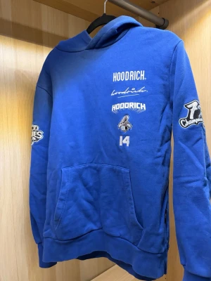 Blå hoodie från Hoodrich - Blå hoodie från Hoodrich med flera tryck och broderade detaljer på bröstet och ärmarna, bland annat texten '7x Champions' och siffran 14. Stor känguruficka framtill och huva. Perfekt för dig som gillar streetwear och sportig stil.