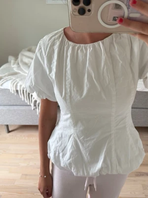 Vit puffärmad blus med resår - Säljer en söt vit blus med puffiga korta ärmar och resår i nederkant. Blusen har en rund halsringning och är tillverkad i ett lätt, krispigt tyg som ger en luftig känsla. Perfekt för en fräsch och stilren look.