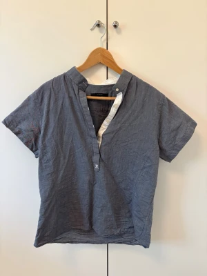 Blå kortärmad henley i linne - Snygg blå henley-skjorta i linne med korta ärmar och ståkrage. Skjortan har en halvknäppning framtill med vita detaljer och en avslappnad passform. Perfekt för dig som gillar en enkel men stilren look.