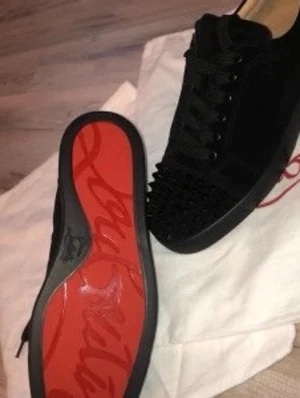 Svarta Louboutin sneakers med nitar - Svarta sneakers från Christian Louboutin med klassisk röd sula och svarta nitar på tån. Skorna har snörning och är tillverkade i mocka och läder. Perfekta för dig som vill sticka ut med en edgy och lyxig stil.