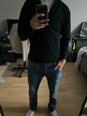 Svart zip-hoodie med huva - En stilren svart hoodie med dragkedja och huva. Hoodien har två fickor framtill och ribbade muddar vid ärmslut och nederkant. Perfekt för en avslappnad och enkel look. Materialet känns mjukt och bekvämt mot huden. Nopprig!