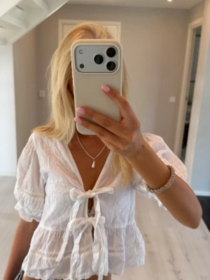 Vit blus med knyt och volang - Söt vit blus med två knyt framtill och volangkant. Blusen har korta puffärmar med spetsdetaljer och v-ringning. Tillverkad i ett lätt bomullstyg som passar perfekt till jeans eller kjol. Superfin till sommaren!