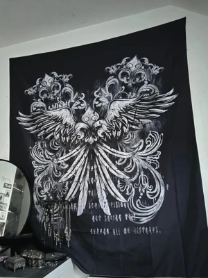 Stor Svart väggbonad - Stor svart väggbonad med detaljerat vitt tryck. med utbredda vingar och ornamentala mönster. Tyget har även text i vitt längst ner. Perfekt som dekoration för att skapa en alternativ eller mystisk vibe i rummet.
