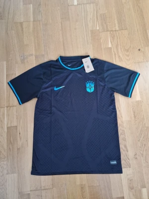 Svart Brasilien Nike fotbollströja - Snygg svart fotbollströja från Nike med Brasilien-emblem och turkosa detaljer. Tröjan har korta ärmar, rund hals och diskret mönster på framsidan. Perfekt för träning eller match. Materialet är lätt och andas bra. Helt nyy!