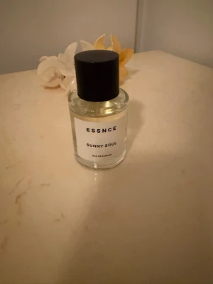 ESSNCE Sunny Soul Eau de Parfum - Parfymflaska i glas med svart rund kork och vit etikett där det står ESSNCE Sunny Soul. Flaskan är genomskinlig och innehåller en ljusgul vätska. Enkel och stilren design som passar perfekt på parfymhyllan.