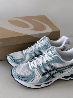 Asics gel kayano 14 - Snygga sneakers från Asics i vitt och ljusblått med svarta detaljer. Ovandelen är i mesh och syntet för bra andningsförmåga och komfort. Klassisk låg modell med vita skosnören och stabil sula. Perfekta för en sportig och trendig look.