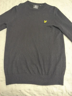 Lyle Scott svart tröja  - Mörkgrå stickad tröja med rund halsringning och ribbade muddar. På bröstet finns en liten gul broderad logga som detalj. Perfekt för lager-på-lager och enkel att matcha med jeans eller kjol.