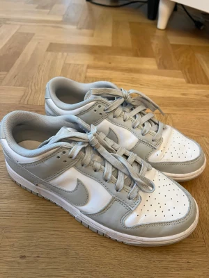 Nike Dunk Low vit och grå - Nike Dunk Low sneakers i vitt och ljusgrått läder med klassisk swoosh på sidan. Lågt skaft, perforerad tåbox och matchande grå skosnören. Stilren och tidlös modell som passar till det mesta. Se skick på bild 