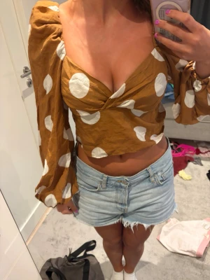 Brun prickig croppad blus - Säljer en brun croppad blus med stora vita prickar och puffiga långärmar. Toppen har omlott-skärning framtill och djup v-ringning som ger en trendig look. Perfekt för dig som vill sticka ut med en unik topp i garderoben.