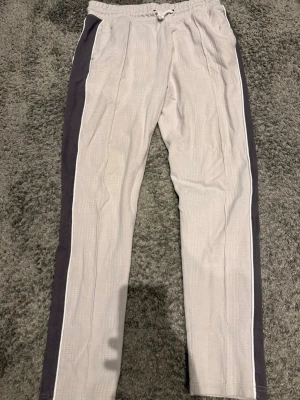 Beige mjukisbyxor med svarta ränder - Säljer ett par beige mjukisbyxor med svarta sidopaneler och vita detaljer längs benen. Byxorna har resår i midjan med dragsko och raka ben. Perfekta för en avslappnad och chill stil.