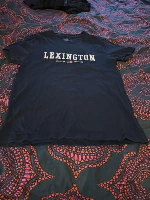 Svart Lexington t-shirt med tryck - Snygg svart t-shirt från Lexington med vit text på bröstet. Klassisk passform och rund halsringning. Tillverkad i mjuk bomull som känns skön mot huden. Perfekt till jeans eller shorts för en avslappnad look.
