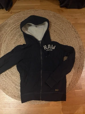 Svart hoodie från G-Star RAW - Säljer en svart hoodie från G-Star RAW med dragkedja och vit text på bröstet. Insidan är fodrad med mjuk teddy för extra komfort. Huva med dragsko och fickor framtill. Perfekt för dig som vill ha en stilren och skön hoodie.