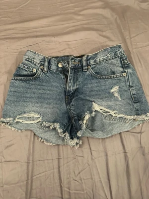 Blå slitna jeansshorts med fransar - Säljer ett par blå jeansshorts med råa, fransiga kanter och slitningar framtill. Klassisk femficksmodell med knapp och dragkedja. Perfekta för sommaren och ger en avslappnad vibe till din outfit.