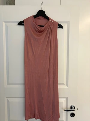 Rosa långklänning från Filippa K - Elegant rosa långklänning från Filippa K i mjukt, slätt material. Klänningen är ärmlös med draperad halsringning och dekorativa knappar på axeln.