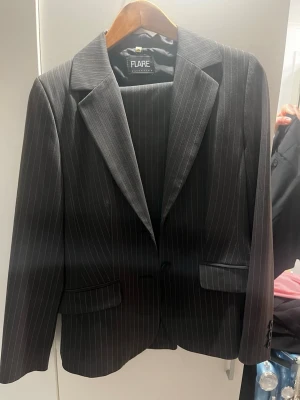 Svart randig kostym från Flare - Snygg svart kostym från Flare med tunna vita ränder. Klassisk kavaj med slag och två fickor framtill, matchande byxor med samma mönster. Perfekt för en stilren look. Aldrig använd
