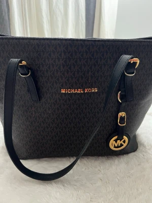 Svart brun  Michael Kors handväska - Snygg svart handväska från Michael Kors med guldiga detaljer och MK-logga. dragkedja. Perfekt för dig som gillar stilrena accessoarer. (Replika) (väskan har en liten vid fläck se andra bilden)