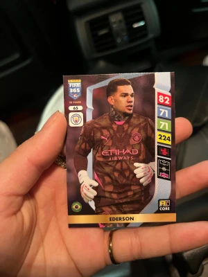 FIFA 365 samlarkort Ederson - Samlarkort från FIFA 365-serien med fotbollsspelaren Ederson. Kortet har färgstark design med bruna, rosa och vita toner samt statistik och klubbmärke. Perfekt för dig som gillar fotboll och vill utöka din samling med en unik spelare.