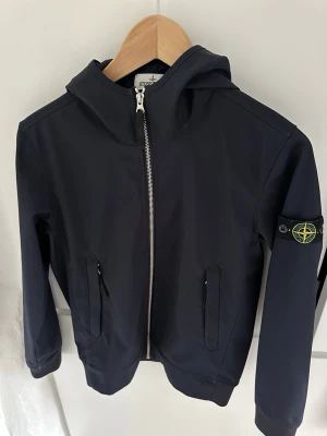 Blå vindjacka från Stone Island - Svart vindjacka från Stone Island med huva och dragkedja framtill. Jackan har två snedställda fickor med lock och den klassiska Stone Island-loggan på vänster ärm. Materialet är slätt och lätt, perfekt för blåsiga dagar.