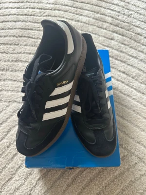Adidas Samba svartvita sneakers - Klassiska Adidas Samba sneakers i svart läder med vita detaljer och bruna gummisulor. Skorna har de ikoniska tre vita ränderna på sidan och texten 'SAMBA' i guld. Lågt skaft och snörning framtill. Perfekta för en sportig och stilren look.