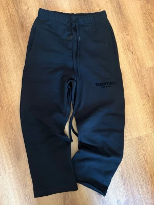 Svarta Essentials mjukisbyxor - Säljer ett par svarta mjukisbyxor från Fear of God Essentials. Byxorna har dragsko i midjan, sidofickor och logga tryckt på benet. Sköna och stilrena, perfekta för en avslappnad look. Materialet känns mjukt och bekvämt. Storlek xxs men passar mer xs. Knappt använda och finns även hoodie 