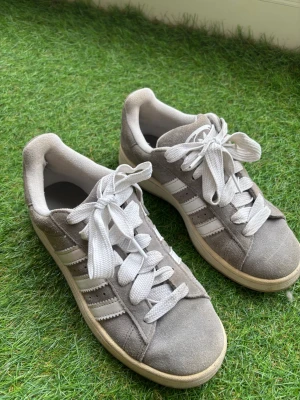 Gråa Adidas Campus 00s i mocka - Säljer ett par gråa Adidas Campus 00s i mocka med klassiska vita ränder på sidorna och vita skosnören. Skorna har en låg profil och en beige sula som ger en retro känsla. Använda under en sommar, och i bra skick!