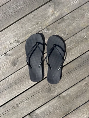 Svarta flip-flops i gummi - Säljer ett par klassiska svarta flip-flops med tunna remmar och räfflad sula. Tillverkade i lätt och flexibelt gummi, perfekta för stranden eller lata sommardagar. Enkel och stilren design som passar till det mesta.