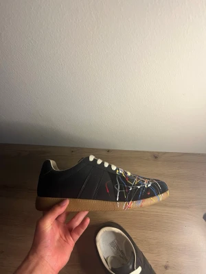 Svarta sneakers med färgstänk - Unika svarta sneakers med vita skosnören och färgstänk i blått, gult, rött och vitt över tåpartiet. Skorna har en klassisk låg siluett och brun sula. Materialet ser ut att vara läder eller konstläder, vilket ger en stilren men kreativ look.