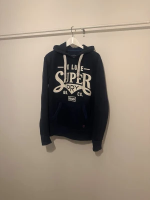 Mörkblå Superdry hoodie med tryck - Mörkblå hoodie från Superdry med stort vitt tryck på bröstet och vita snören, storleken är xs men jag skulle säga att den sitter som S, jag är 175 60kg och den är lite stor. Mycket bra skick men trycket på bröstet är lite slitet annars dunder, hör av dig vid minsta lilla mvh Max 