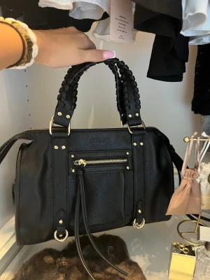 Svart handväska med flätade handtag - Snygg svart handväska i konstläder med flätade handtag och guldfärgade detaljer. Väskan har en framficka med dragkedja och dekorativa remmar. Perfekt storlek för vardagsprylar och ger en cool touch till din outfit. Köptes för 899