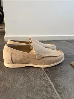 Beige mocka loafers - Snygga loafers i beige mocka med klassisk design och rundad tå. Skorna har diskreta sömmar och en låg, ljus sula som ger en stilren look. Perfekta för dig som gillar en clean och enkel stil. Materialet är mjukt och ger en exklusiv känsla.