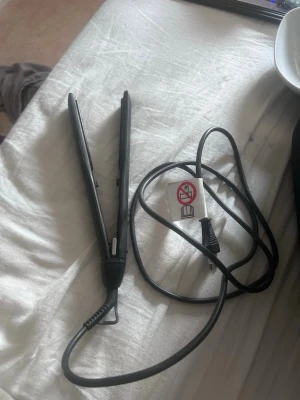 Svart plattång för styling - En svart plattång med sladd, perfekt för att styla och räta ut håret. Plattången har en smal och avlång design med släta plattor och enkel knapp på sidan. Passar dig som vill fixa frisyren snabbt och smidigt.