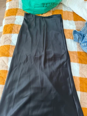 Svart långkjol i satin från Zara - Snygg svart långkjol från Zara i mjukt satinliknande material. Kjolen har en enkel och stilren design med rak passform som ger en elegant look. Perfekt att styla med både sneakers och boots för en trendig vibe.