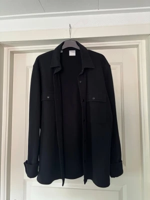 Svart overshirt med knappar - Stilren svart overshirt med klassisk krage och tryckknappar framtill. Två stora bröstfickor med lock och långa ärmar med manschetter. Perfekt att bära som lager över t-shirt eller hoodie för en avslappnad look.
