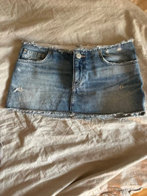 Kort jeanskjol med råa kanter - Säljer en kort jeanskjol i blå denim med slitna detaljer och råa kanter. Kjolen har klassiska fickor fram och bak samt knapp och dragkedja framtill. Perfekt för dig som gillar en edgy och avslappnad stil.