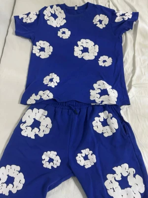 Blått set med vita molntryck - Säljer ett blått set med t-shirt och shorts, dekorerat med stora vita molnliknande tryck över hela plaggen. Setet har avslappnad passform och elastisk midja på shortsen. Perfekt för en chill och lekfull stil. Storlek L men passar mer som M