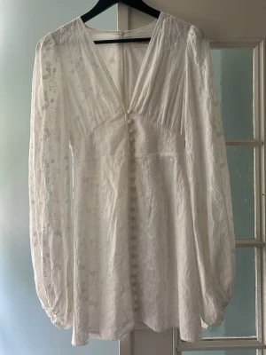 Adoore. Vit modern långärmad klänning - Romantisk vit klänning med blommigt mönster, v-ringning och långa puffärmar. Klänningen har dekorativa knappar framtill och är sydd i ett lätt, luftigt tyg som ger en elegant känsla. Perfekt för sommarens alla tillfällen.