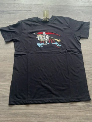 Svart Burberry t-shirt med tryck - Snygg svart t-shirt från Burberry med färgglatt tryck av riddare på häst framtill. Klassisk passform och rund halsringning. Tillverkad i mjuk bomull som känns skön mot huden. Perfekt för dig som vill ha en stilren men ändå unik look.