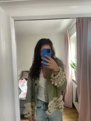 Grön jacka  - Snygg grön overshirt med dekorativa fransar och spetsdetaljer i beige vid ärmslut och nederkant. Jackan har knappar framtill, två bröstfickor och en loose passform. Perfekt för dig som vill sticka ut med en unik och trendig look.