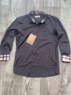 Svart skjorta från Burberry - Svart skjorta från Burberry med klassiska rutiga detaljer på insidan av kragen, knappslån och ärmsluten. Skjortan har knappar framtill och lång ärm. Perfekt för dig som gillar stilrena plagg med exklusiv känsla.