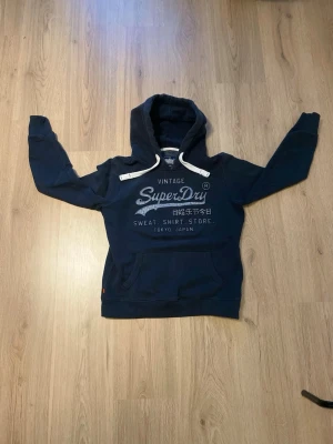Superdry Hoodie 👕 - Superdry hoodie 🔥 Mörkblå hoodie från Superdry med klassiskt logotryck framtill. Mjuk och bekväm modell med huva och känguruficka – perfekt för en avslappnad vardagslook. I fint begagnat skick. 📏 Storlek: S 📦 Skick: Bra begagnat skick 💬 Hör gärna av dig vid frågor eller om du vill ha fler bilder!