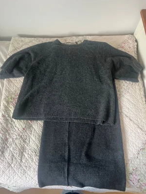 Mörkgrått stickat set med kjol - Säljer ett mörkgrått stickat set med en oversized tröja med halvlånga ärmar och en matchande rak kjol. Setet är mjukt och bekvämt, perfekt för dig som gillar en enkel men stilren look. Materialet känns som ullblandning. Settet är helt oanvänd och i storlek xs/s 