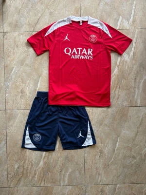 PSG fotbollströja och shorts set - Säljer ett PSG matchställ med röd kortärmad tröja och mörkblå shorts. Tröjan har vita detaljer på axlarna, PSG-logga och Jordan-logga samt Qatar Airways-tryck. Shortsen är mörkblå med vita detaljer och PSG-logga. Perfekt för fotbollsträning eller match.