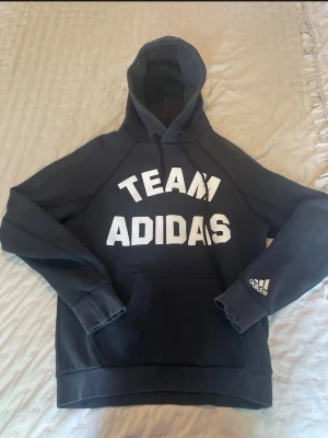 Svart hoodie Team Adidas - Snygg svart hoodie från Adidas med stor vit 'TEAM ADIDAS'-text på bröstet och Adidas-logga på ärmen. Klassisk huva med dragsko och stor magficka framtill. Perfekt för en sportig och avslappnad stil.
