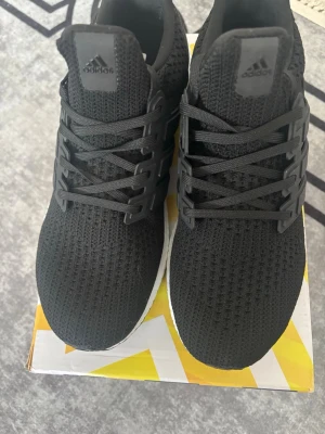 Svarta Adidas Ultraboost sneakers - Säljer ett par helsvarta Adidas Ultraboost sneakers med stickad ovandel och klassisk Boost-sula. Skorna har svarta snören och en flexibel, ventilerande meshstruktur. Yttersulan är i slitstarkt gummi från Continental för extra grepp.