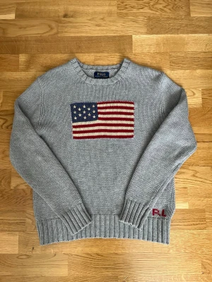 Ralph Lauren  - Snygg grå stickad tröja från Polo Ralph Lauren med en stor amerikansk flagga på bröstet och diskret röd 'P.L.' brodyr vid nederkanten. Tröjan har rund halsringning, långa ärmar och ribbade muddar. Perfekt för dig som gillar klassisk och avslappnad stil.