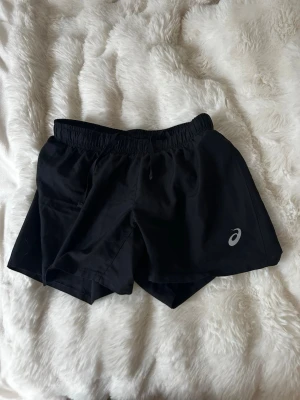 Svarta träningsshorts från Asics - Säljer ett par svarta träningsshorts från Asics med elastisk midja och snörning. Shortsen har en enkel design med Asics-logga på ena benet och är perfekta för sport eller gym. Lätta och bekväma att röra sig i.