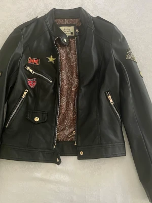 Svart skinnjacka med patches - Svart skinnjacka med flera snygga patches och dragkedjor i guld. Jackan har tryckknapp och paisleymönstrat innerfoder. 