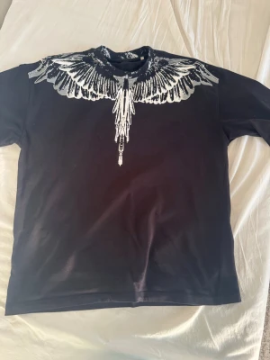 Svart t-shirt med vitt fjädertryck - Säljer en svart t-shirt med ett fett vitt fjädermönster runt halsen, både fram och bak. T-shirten har klassisk passform och korta ärmar. Perfekt för dig som vill sticka ut med en unik design. Storlek XL men passar som L
