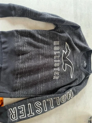 Hollister sweatshirt - Grå  sweatshirt från Hollister med vit logga och text på bröstet samt ärmen. Tröjan har rund halsringning och ribbade muddar. Materialet är mjukt och bekvämt, perfekt för en avslappnad stil.